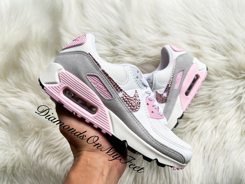 May include: White and grey Nike Air Max 90 sneakers with pink accents and rhinestones on the swoosh logo. The shoes have a pink air bubble on the heel and a white midsole.
