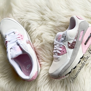 May include: White and pink Nike Air Max 90 sneakers with a pink and white swoosh logo and rhinestones on the swoosh. The sneakers have a white midsole and a pink outsole. The text "Diamonds On My Feet" is visible on the right shoe.