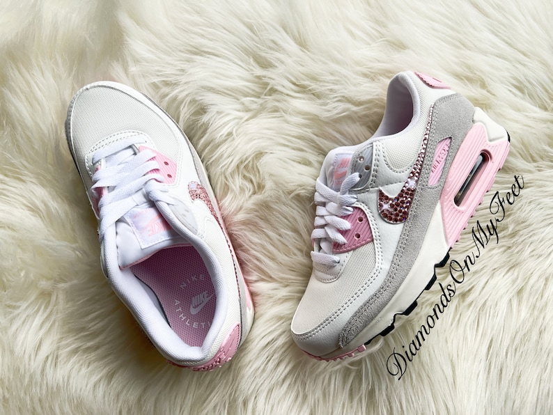 May include: White and pink Nike Air Max 90 sneakers with a pink and white swoosh logo and rhinestones on the swoosh. The sneakers have a white midsole and a pink outsole. The text "Diamonds On My Feet" is visible on the right shoe.