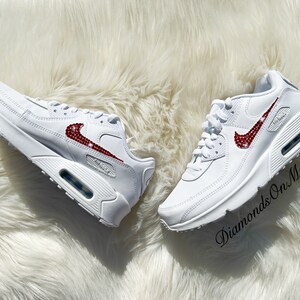 May include: White leather sneakers with a red rhinestone Nike swoosh. The shoes are on a white fluffy surface. The text "Diamonds On My Feet" is visible in the lower right corner of the image.
