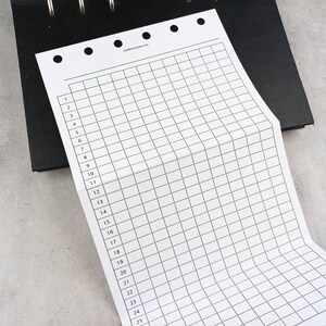May include: A white, grid-lined paper sheet with numbered rows and columns, partially covering a black binder. The paper has a series of small holes along the top edge. The grid is designed for organizational purposes.