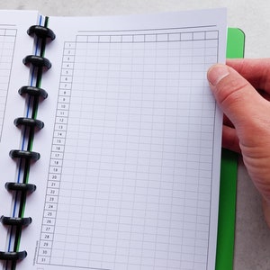 Puede incluir: Un cuaderno blanco con una encuadernaci&oacute;n en espiral negra y un patr&oacute;n de cuadr&iacute;cula en las p&aacute;ginas. El cuaderno est&aacute; abierto en una p&aacute;gina con los n&uacute;meros del 1 al 31 listados en el lateral.