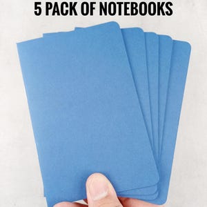 May include: A hand holding a pack of five blue notebooks. The notebooks are a solid light blue colour and have rounded corners. The text "5 PACK OF NOTEBOOKS" is printed in black at the top of the image.