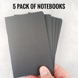 May include: A hand holding a stack of five black notebooks. The notebooks are rectangular with rounded corners. The text "5 PACK OF NOTEBOOKS" is displayed in black, centered at the top of the image.