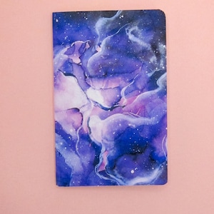 May include: A notebook with a galaxy-inspired design. The cover features a swirling pattern of purple, blue, and white, resembling a nebula. The background is a dark blue with white speckles, representing stars.