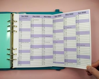 May include: A close-up of a planner with a turquoise cover and gold rings. The planner is open to the months of January to June 2022. Each month is displayed on a separate page with a light purple background and a white grid. The days of the week are labelled with the abbreviations S, M, T, W, T, F, and S. Some days have special holidays marked, such as New Year's Day, Groundhog Day, Valentine's Day, President's Day, Mother's Day, Father's Day, and Memorial Day.
