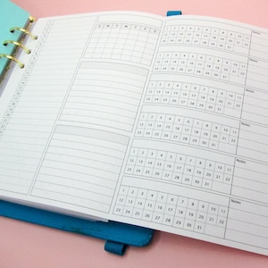 Peut inclure: Un agenda blanc avec une couverture bleue et des anneaux dorés. L'agenda a une vue mensuelle du calendrier et une section de liste de tâches quotidiennes. L'agenda est ouvert à une page avec le mois de janvier.