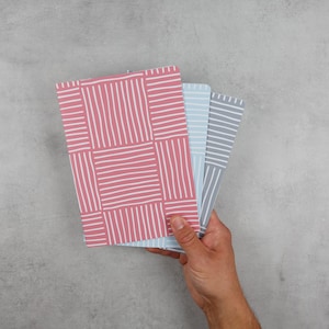 May include: Three notebooks with different geometric patterns. The notebooks are pink, light blue, and grey. The notebooks are stacked on top of each other.