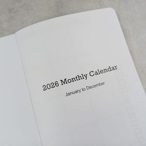 May include: Open notebook with the text "2026 Monthly Calendar" printed in black. The calendar spans from January to December. The pages are white, and the background is a light grey.