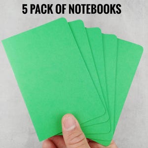 May include: A hand holding a stack of five bright green notebooks. The notebooks are rectangular with rounded corners. The text "5 PACK OF NOTEBOOKS" is printed in black at the top of the image.