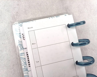 Può includere: Primo piano di un'agenda con una copertina in plastica trasparente. Le pagine bianche hanno un layout a griglia e la parola "Mese" in alto. L'agenda ha anelli di plastica blu e linguette con "Luglio" e "Febbraio".
