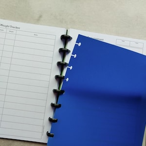 Puede incluir: Una libreta azul con una espiral negra. La portada tiene el texto "Height & Weight Tracker" y columnas para la fecha, la altura, el peso y las notas. La contraportada es azul y tiene el texto "Appointment" y columnas para la fecha y la hora.
