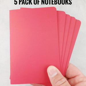 May include: A hand holding a stack of five red notebooks. The notebooks are rectangular with rounded corners. The text "5 PACK OF NOTEBOOKS" is printed in black at the top of the image.