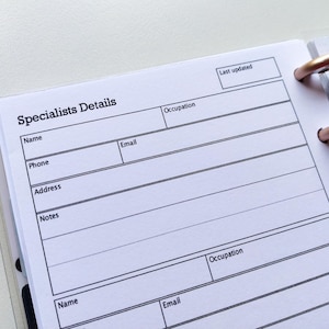 May include: A white page with black text and lines for filling in contact information. The page is titled "Specialists Details" and includes fields for name, phone, email, address, notes, and occupation. The page is part of a bound notebook with gold rings.