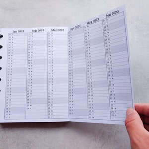 Puede incluir: Un calendario en blanco y negro con encuadernaci&oacute;n en espiral. El calendario est&aacute; abierto en junio de 2023, con los meses de enero a mayo visibles en el fondo.