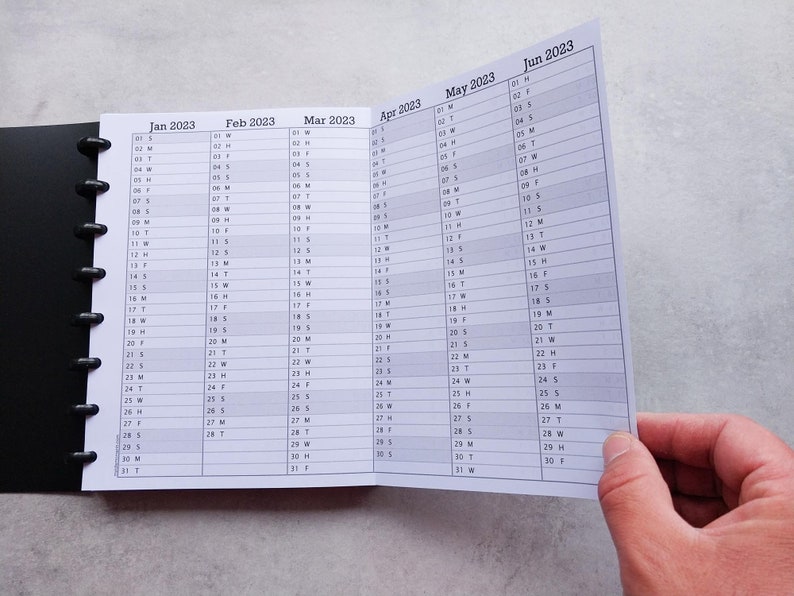 Puede incluir: Un calendario en blanco y negro con encuadernaci&oacute;n en espiral. El calendario est&aacute; abierto en junio de 2023, con los meses de enero a mayo visibles en el fondo.