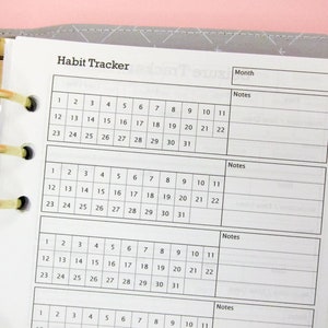 May include: A white habit tracker insert with a gold ring binder. The habit tracker has a grid layout with numbered days and space for notes. The text "Habit Tracker" is at the top.