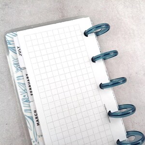 Pode incluir: Um planejador com uma página quadriculada e anéis de plástico azuis. O planejador tem divisórias mensais com os meses "Julho", "Fevereiro" e "Março". As divisórias têm um design floral azul. O planejador está aberto, mostrando o papel quadriculado.