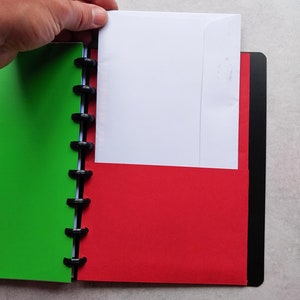 Puede incluir: Un cuaderno verde y rojo con un encuadernador negro y un sobre blanco que sobresale del lado rojo.