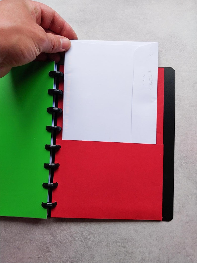 Puede incluir: Un cuaderno verde y rojo con un encuadernador negro y un sobre blanco que sobresale del lado rojo.