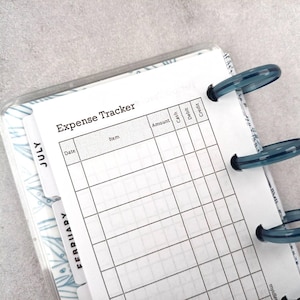 May include: An open planner with an expense tracker page visible. The page is white with black grid lines and the title "Expense Tracker". The planner has blue plastic rings and dividers labeled "July" and "February".