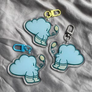 May include: Three acrylic keychains featuring a cartoon cloud character in light blue with a yellow balloon. Each keychain has a different colored clasp: yellow, silver, and blue. The cloud character has a sad expression and is wearing light blue boots.