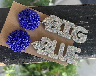 May include: A pair of earrings featuring blue beaded pom-poms and silver glitter letters spelling "BIG BLUE".