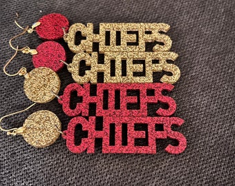 May include: Glittery red and gold "Chiefs" earrings. The earrings have a circular stud and a word-shaped pendant. The word "Chiefs" is spelled out in a bold, blocky font. The earrings are designed for sports fans.