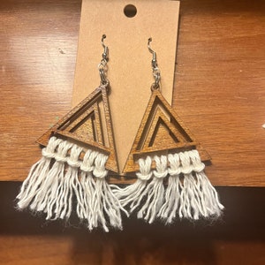 May include: A pair of wooden triangle earrings with white fringe tassels. The earrings are made of light brown wood and have a geometric design.