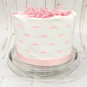 May include: A white fabric gift basket with a pink crown pattern and a pink ribbon. The basket is filled with pink tissue paper and is sitting on a clear glass cake stand.