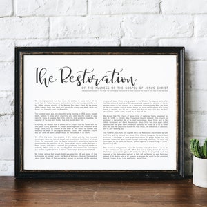 May include: A framed print with the title "The Restoration" in elegant script. The print features a black frame and contains text detailing the restoration of the Gospel of Jesus Christ. The print is displayed on a wooden surface with two potted cacti.