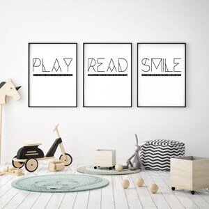 May include: Three black framed prints with white backgrounds and black text. The prints read "PLAY", "READ", and "SMILE". The prints are hanging on a white wall in a room with a wooden rocking horse, a wooden scooter, a wooden storage bin, and a gray and white striped beanbag chair.