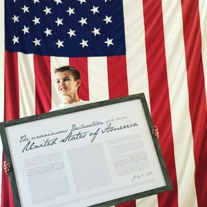 May include: A framed copy of the Declaration of Independence, with the text "The unanimous Declaration of the thirteen United States of America" and the date "July 4, 1776" visible. A young person is holding the framed document in front of an American flag.