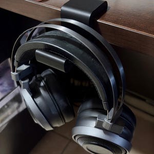 May include: Black over-ear headphones hanging from a black headphone stand. The headphones have a padded headband and earcups, with silver accents. The stand is attached to a wooden surface.