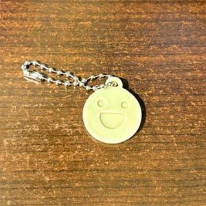 May include: A light yellow keychain charm with a smiling face design. The charm is round with a small loop at the top, attached to a silver ball chain. The background is a wooden surface.