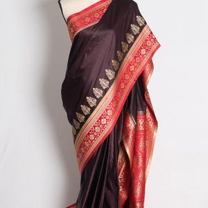 May include: A dark brown saree featuring a wide, vibrant red border adorned with intricate gold metallic floral patterns. The fabric has a smooth, satin-like sheen, and the border includes a repeating gold motif along the edge.