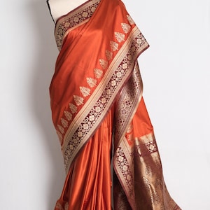 May include: A vibrant burnt orange saree featuring a wide, deep maroon border with intricate gold metallic floral and paisley patterns. The fabric has a smooth, lustrous finish, and the decorative border extends along the edges and the pallu.