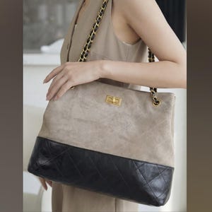 Pode incluir: Uma bolsa tote de duas cores com uma parte superior de camur&ccedil;a bege clara e uma base de couro acolchoado preto. A bolsa apresenta um fecho dourado e uma al&ccedil;a de corrente, segurada por uma pessoa.