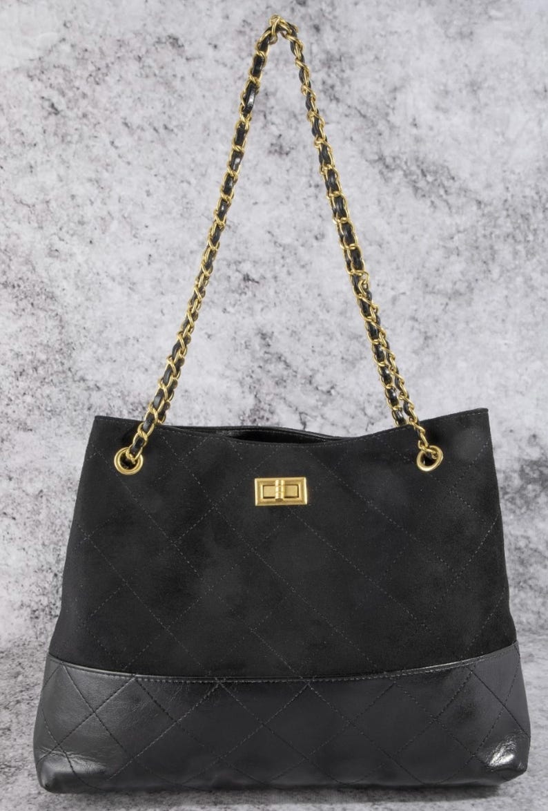 Pode incluir: Bolsa tote preta com al&ccedil;a de corrente dourada. A bolsa apresenta uma parte superior de camur&ccedil;a acolchoada e uma base de couro com costura de diamante. Um fecho retangular dourado est&aacute; centrado na frente.