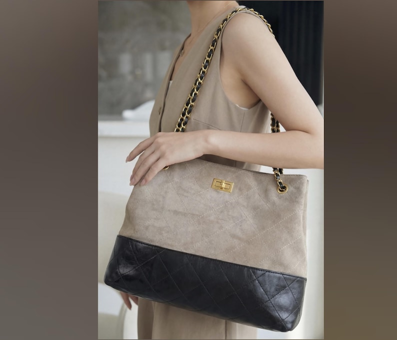 Pode incluir: Uma bolsa tote de duas cores com uma parte superior de camur&ccedil;a bege clara e uma base de couro acolchoado preto. A bolsa apresenta um fecho dourado e uma al&ccedil;a de corrente, segurada por uma pessoa.