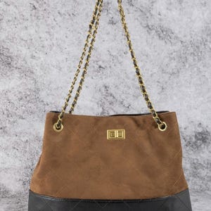 Pode incluir: Bolsa tote de camur&ccedil;a marrom com fundo de couro preto e al&ccedil;a de corrente dourada. A bolsa tem um padr&atilde;o acolchoado na camur&ccedil;a e um fecho dourado. Um acess&oacute;rio elegante.
