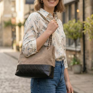 Pode incluir: Uma bolsa tote de couro acolchoada bege e preta com uma al&ccedil;a de corrente dourada. A bolsa &eacute; carregada por uma pessoa que usa uma blusa com estampa floral e jeans azuis. O fundo &eacute; uma rua de paralelep&iacute;pedos.