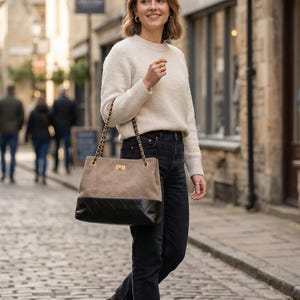 Pode incluir: Uma mulher com um su&eacute;ter creme e jeans pretos, carregando uma bolsa de duas cores com uma al&ccedil;a de corrente dourada. A bolsa tem uma se&ccedil;&atilde;o superior bege e uma se&ccedil;&atilde;o inferior preta. Ela est&aacute; andando em uma rua de paralelep&iacute;pedos.