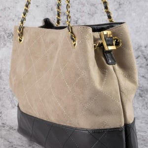 Pode incluir: Bolsa tote de camur&ccedil;a bege com base de couro preta. A bolsa apresenta um padr&atilde;o acolchoado em losangos e al&ccedil;as de corrente douradas. Um fecho de couro preto e dourado prende a parte superior.
