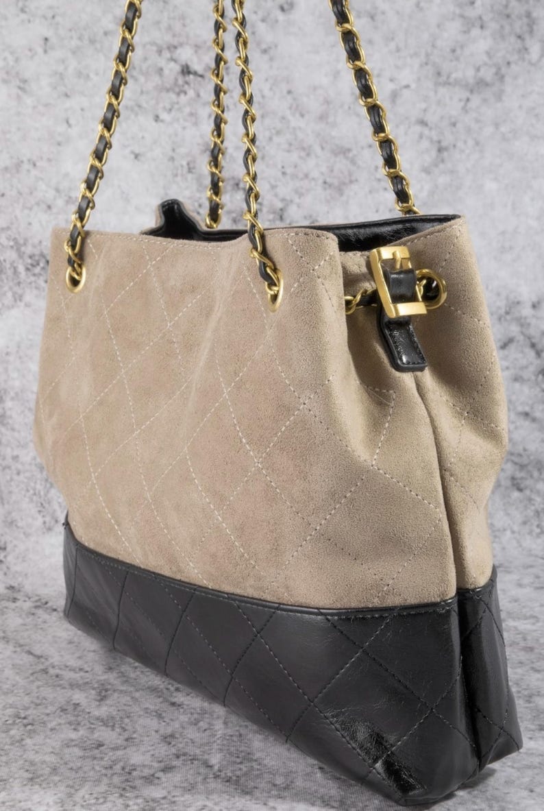 Pode incluir: Bolsa tote de camur&ccedil;a bege com base de couro preta. A bolsa apresenta um padr&atilde;o acolchoado em losangos e al&ccedil;as de corrente douradas. Um fecho de couro preto e dourado prende a parte superior.