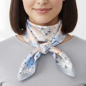 May include: A square silk scarf with a floral pattern in white, light blue, and peach. The scarf is tied around the neck, showcasing the delicate design and soft color palette. The scarf is a fashion accessory.