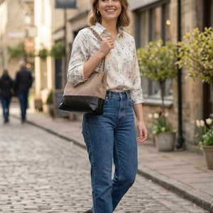 Pode incluir: Uma pessoa caminha por uma rua de paralelep&iacute;pedos, usando uma blusa com estampa floral, jeans azuis e botas pretas. Ela carrega uma bolsa tote bege e preta com uma al&ccedil;a de corrente. A placa ao fundo diz "ARTISAN CHRATS".