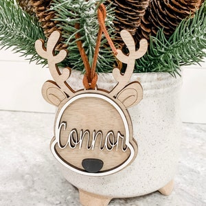 May include: A wooden reindeer ornament with a brown suede cord for hanging. The ornament has a white outline and the name "Connor" is written in white inside the outline. The ornament is sitting in a white ceramic pot with a pine tree branch.