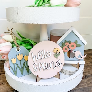 May include: A white tiered tray with a pink and white "hello spring" sign, a blue birdhouse with a bird, and a blue tag with yellow tulips. The tray is decorated with spring flowers.