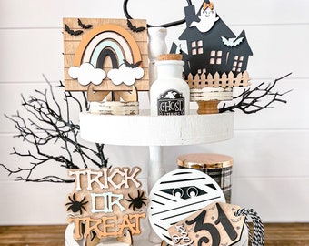 Könnte beinhalten: Ein zweistufiges weißes Tablett mit Halloween-Motiven aus Holz. Die Schilder zeigen einen Regenbogen, eine Mumie, ein Gespenst, eine Spinne und die Worte "Trick or Treat". Das Tablett enthält auch ein Glasgefäß mit dem Etikett "Ghost Tears" und ein schwarz-weiß gestreiftes Band.